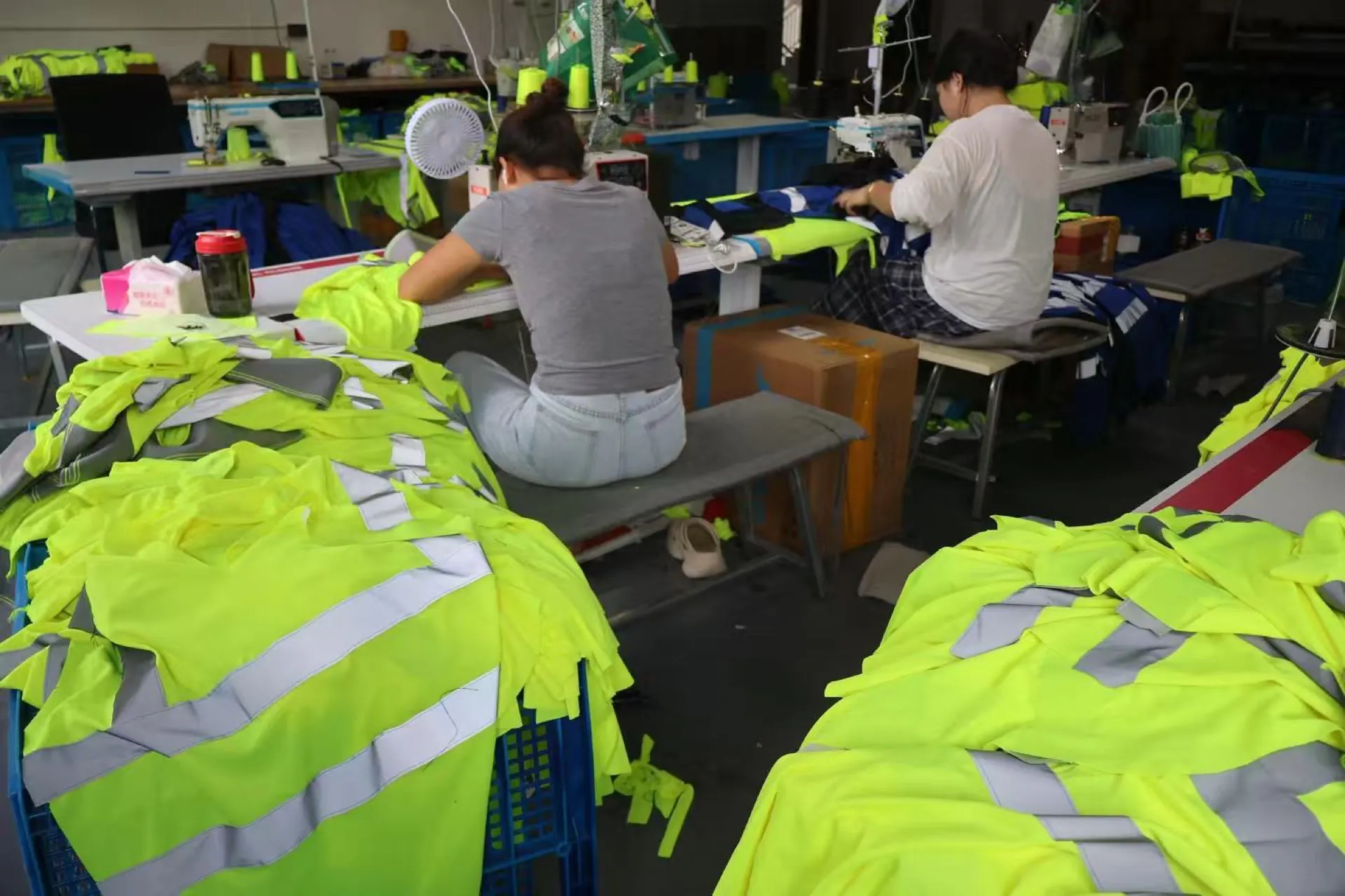 Two garment workers handling fluorescent reflective workwear at sewing stations